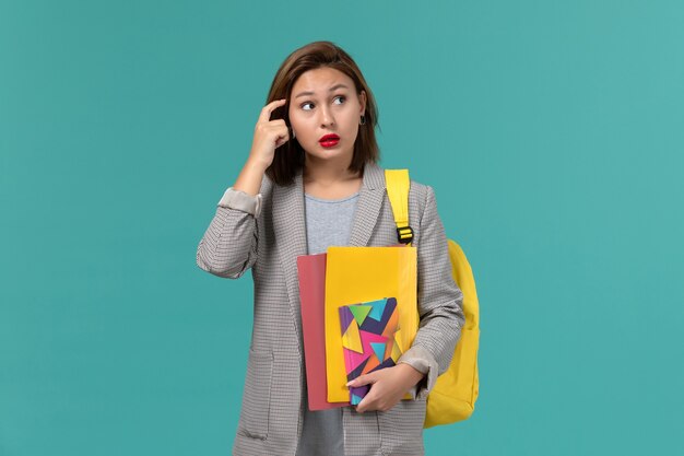 Vue de face de l'étudiante en veste grise portant un sac à dos jaune tenant des fichiers et un cahier amincissant sur le mur bleu