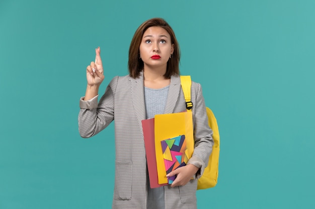 Vue de face de l'étudiante en veste grise portant un sac à dos jaune tenant des fichiers et un cahier croisant ses doigts sur le mur bleu