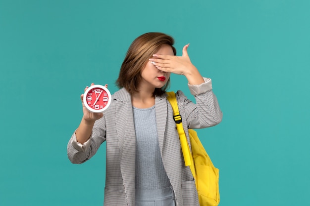 Vue de face de l'étudiante en veste grise portant un sac à dos jaune tenant des horloges et couvrant son visage sur un mur bleu