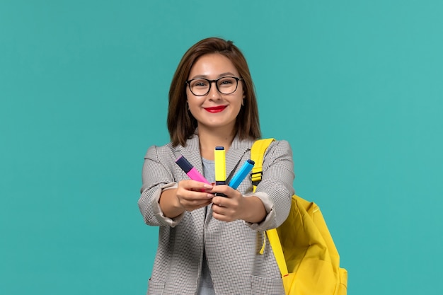 Vue de face de l'étudiante en veste grise portant son sac à dos jaune tenant des feutres sur le mur bleu clair