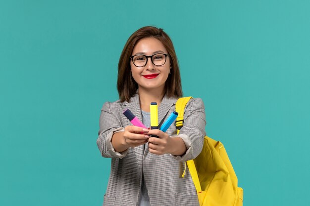 Vue de face de l'étudiante en veste grise portant son sac à dos jaune tenant des feutres sur le mur bleu clair