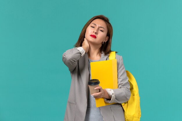 Vue de face de l'étudiante en veste grise portant son sac à dos jaune tenant des fichiers et du café sur le mur bleu