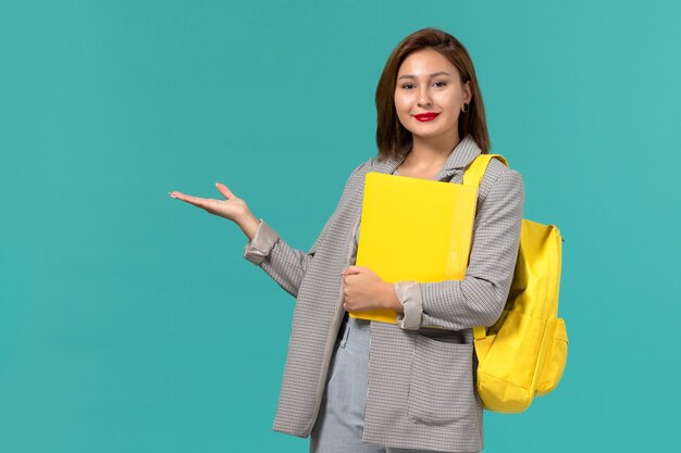 Vue de face de l'étudiante en veste grise portant son sac à dos jaune et tenant des fichiers sur le mur bleu clair