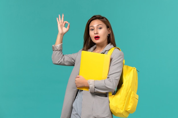 Vue de face de l'étudiante en veste grise portant son sac à dos jaune et tenant des fichiers sur le mur bleu clair