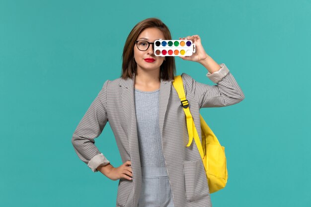 Vue de face de l'étudiante en veste grise portant son sac à dos jaune tenant des peintures pour dessiner sur un mur bleu clair