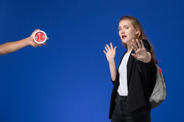 Vue de face étudiante en veste noire portant un sac à dos sur le mur bleu