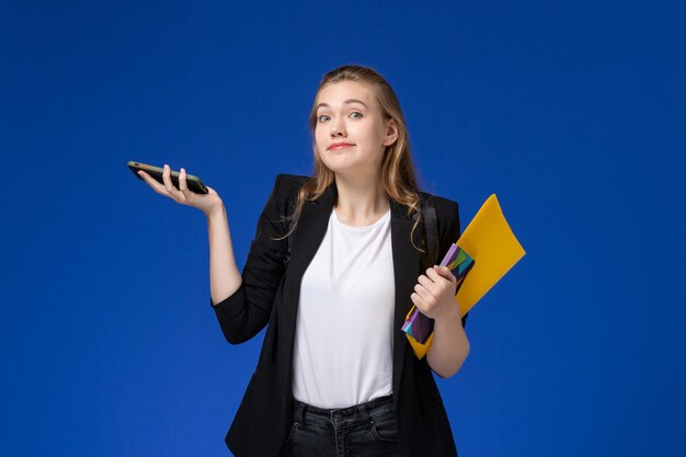 Vue de face étudiante en veste noire portant un sac à dos tenant le fichier et un cahier avec téléphone sur les livres de leçon de l'université collège mur bleu