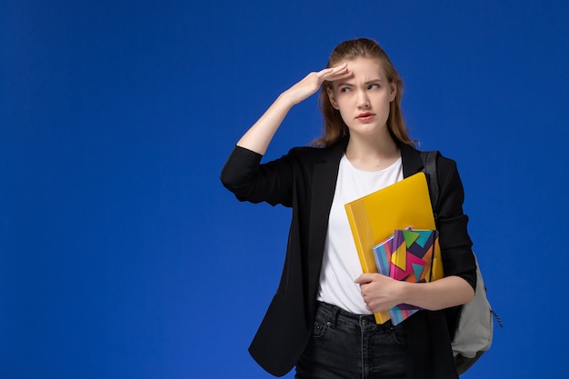 Vue de face étudiante en veste noire portant sac à dos tenant le fichier avec des cahiers de réflexion sur le mur bleu leçon d'université college