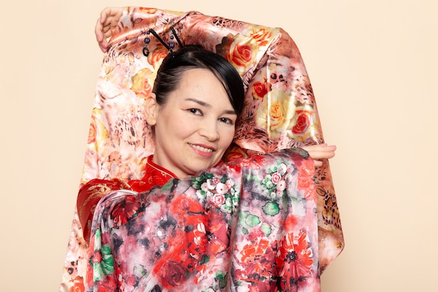 Une vue de face exquise geisha japonaise en robe japonaise rouge traditionnelle posant avec un tissu conçu avec des fleurs élégant souriant sur la cérémonie de fond crème Japon