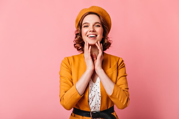 Vue de face de la femme adorable en riant en béret jaune. Photo de Studio de jeune fille souriante aux cheveux ondulés posant sur fond rose.