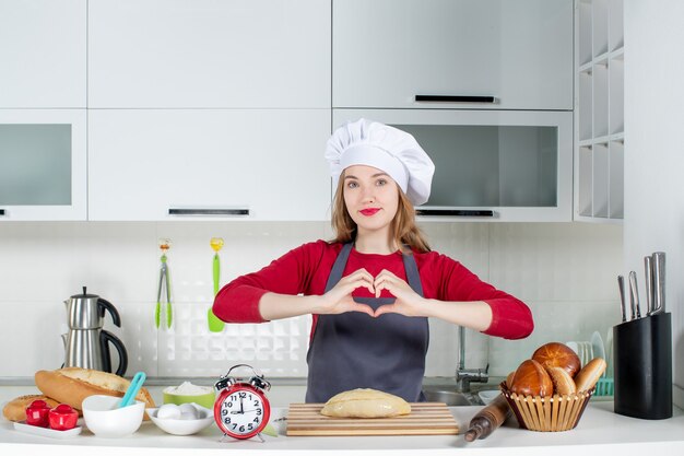 Vue de face femme blonde en chapeau de cuisinier et tablier faisant signe de coeur dans la cuisine