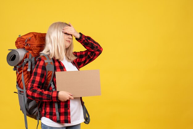 Vue de face femme blonde avec son sac à dos mettant la main sur ses yeux tenant carton