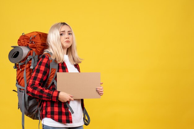 Vue de face femme blonde voyageur avec son sac à dos tenant carton sur mur jaune