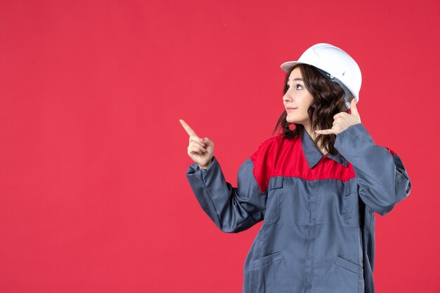 Vue de face d'une femme builder curieuse en uniforme avec un casque et faisant un geste de l'appel vers le haut sur fond rouge isolé