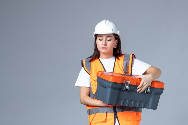 Vue de face d'une femme builder tenant une boîte à outils sur un mur gris