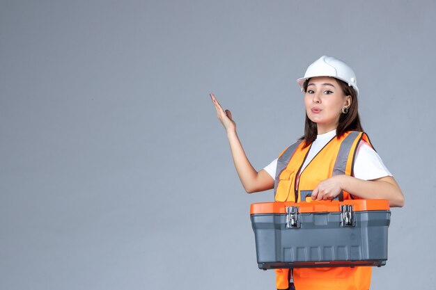 Vue de face d'une femme builder tenant une boîte à outils sur un mur gris