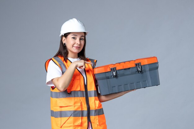 Vue de face d'une femme builder tenant une boîte à outils sur un mur gris