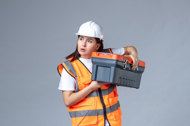 Vue de face d'une femme builder tenant une boîte à outils sur un mur gris