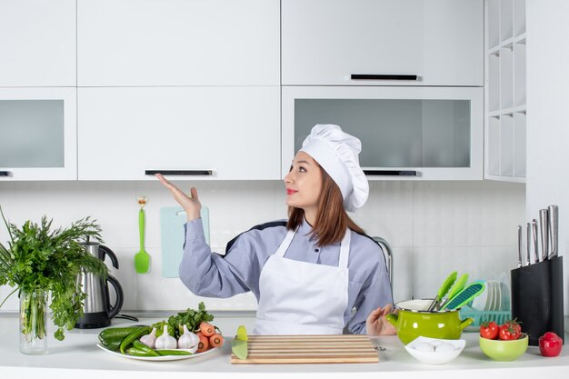 Vue de face d'une femme chef positive et de légumes frais pointant quelque chose sur le côté droit dans la cuisine blanche