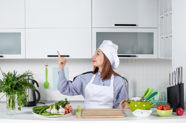 Vue de face d'une femme chef souriante et positive et de légumes frais pointant vers le côté droit dans la cuisine blanche