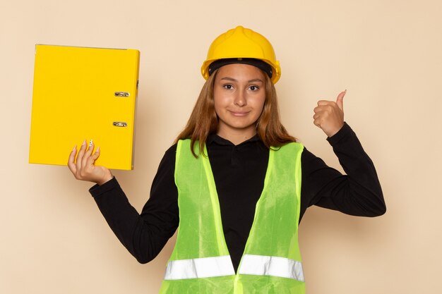 Vue de face femme constructeur en casque jaune tenant un document jaune souriant sur mur blanc