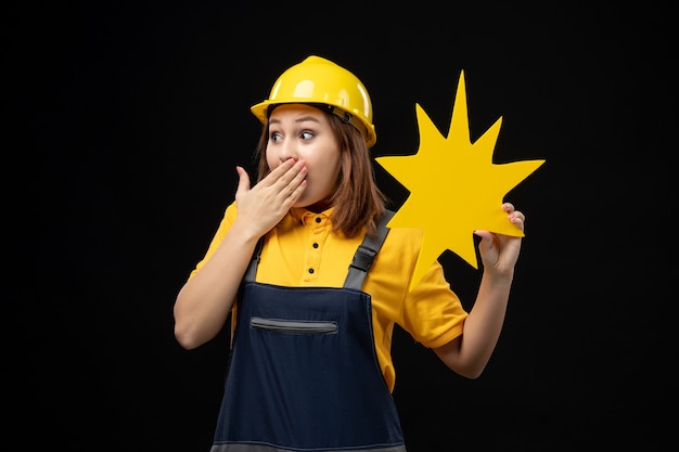 Vue de face femme constructeur en uniforme tenant une figure jaune sur le mur noir