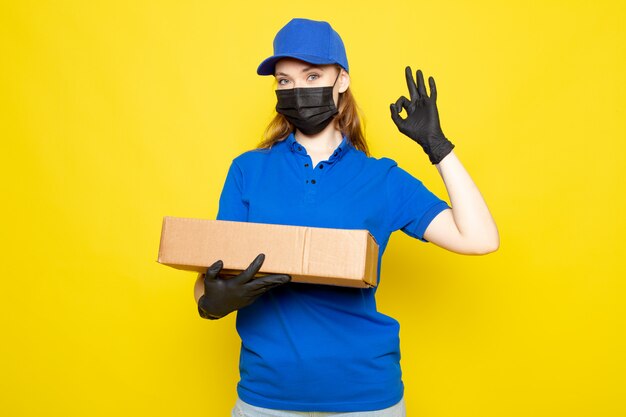 Une vue de face femme courier attrayant en polo bleu capuchon bleu et jeans holding package in black gants masque de protection noir sur le fond jaune job service alimentaire