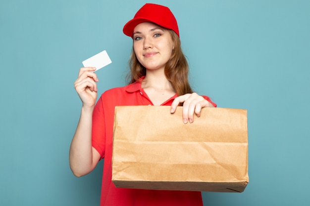 Une vue de face femme courier attrayant en polo rouge casquette rouge et jeans holding package et carte blanche posant souriant sur le fond bleu emploi du service alimentaire