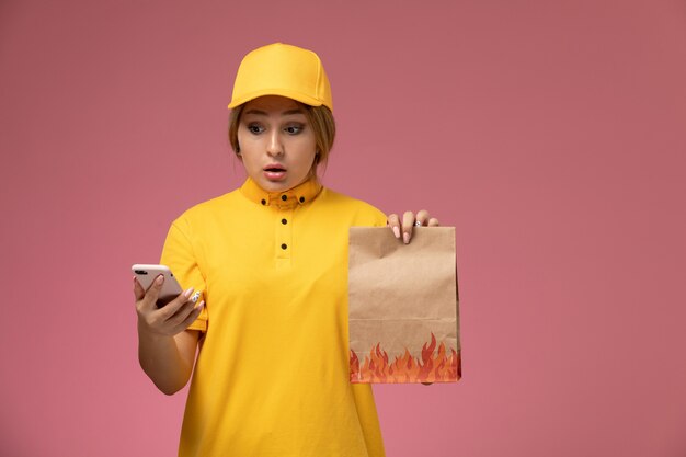 Vue de face femme courrier en cape jaune uniforme jaune tenant le paquet de nourriture et à l'aide de smartphone sur fond rose travail de livraison uniforme travail couleur