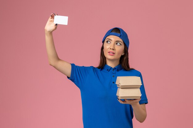 Vue de face femme courrier en cape uniforme bleu holding card et petits colis de livraison sur mur rose clair, travail de livraison des employés de service