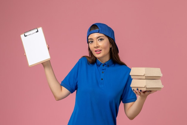 Vue de face femme courrier en cape uniforme bleu tenant le bloc-notes et petits colis de livraison sur le mur rose clair, la livraison des employés de l'emploi de service