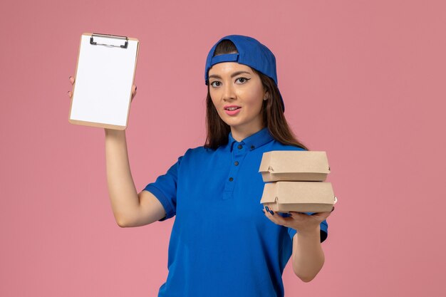 Vue de face femme courrier en cape uniforme bleu tenant le bloc-notes et petits colis de livraison sur le mur rose clair, la livraison des employés de service