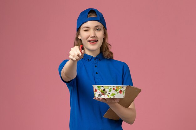 Vue de face femme courrier en cape uniforme bleu tenant le bol de livraison et le bloc-notes sur le mur rose clair, employé de travail de prestation de services