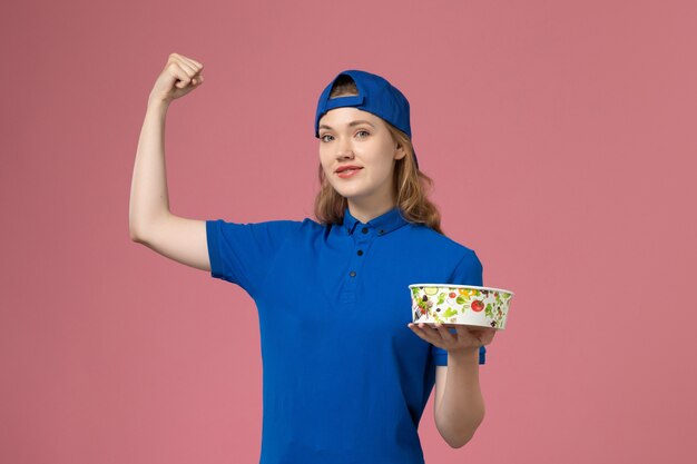 Vue de face femme courrier en cape uniforme bleu tenant le bol de livraison fléchissant sur le mur rose clair, employé de la prestation de services