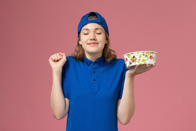 Vue de face femme courrier en cape uniforme bleu tenant le bol de livraison sur le mur rose clair, employé de la prestation de services