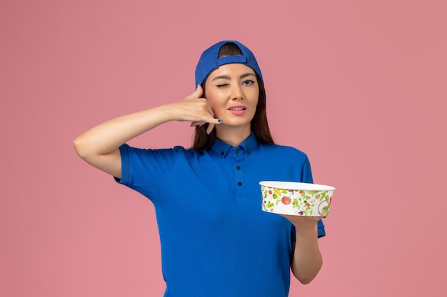 Vue de face femme courrier en cape uniforme bleu tenant le bol de livraison sur le mur rose clair, la livraison des employés de service d'emploi