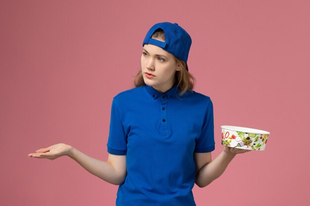 Vue de face femme courrier en cape uniforme bleu tenant le bol de livraison sur le mur rose, employé de la prestation de services des travailleurs