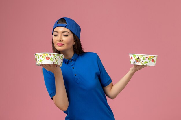 Vue de face femme courrier en cape uniforme bleu tenant des bols de livraison sentant sur le mur rose clair, la livraison des employés de service