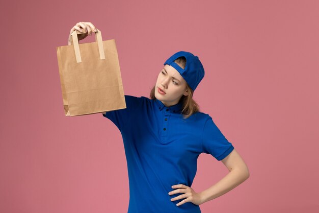 Vue de face femme courrier en cape uniforme bleu tenant le paquet de livraison de papier sur le mur rose, employé de livraison d'emploi de service