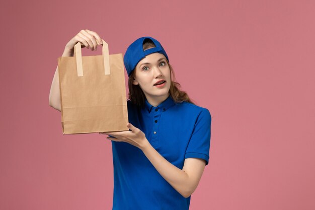 Vue de face femme courrier en cape uniforme bleu tenant le paquet de livraison de papier sur le mur rose, employé de la prestation de services