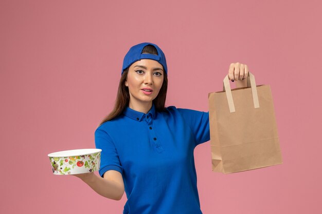 Vue de face femme courrier en cape uniforme bleu tenant le paquet de papier de livraison et bol sur le mur rose, employé de service la prestation de travail