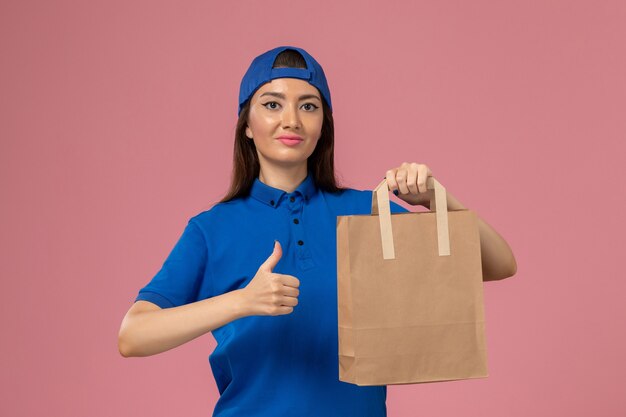 Vue de face femme courrier en cape uniforme bleu tenant le paquet de papier de livraison sur le mur rose, employé de service qui livre du travail