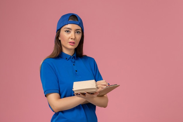 Vue de face femme courrier en cape uniforme bleu tenant un petit paquet de livraison vide avec bloc-notes sur mur rose clair, la livraison de l'entreprise de travail des employés