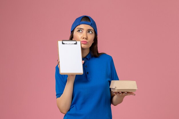 Vue de face femme courrier en cape uniforme bleu tenant un petit paquet de livraison vide avec bloc-notes en pensant au mur rose