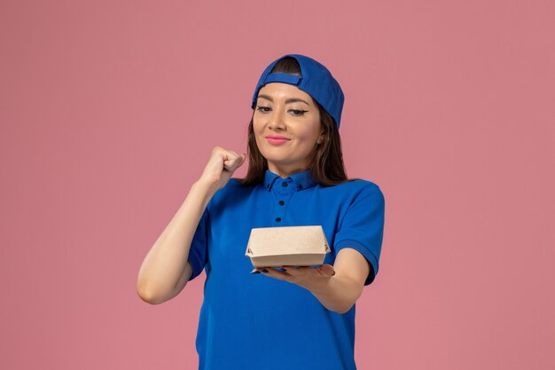 Vue de face femme courrier en cape uniforme bleu tenant un petit paquet de livraison vide et se réjouissant sur le mur rose, la livraison de l'entreprise de services aux employés
