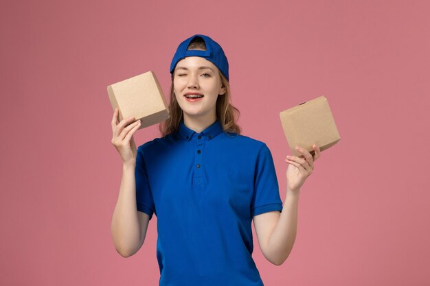 Vue de face femme courrier en cape uniforme bleu tenant peu de colis alimentaires de livraison sur le mur rose, travailleur employé du service de livraison