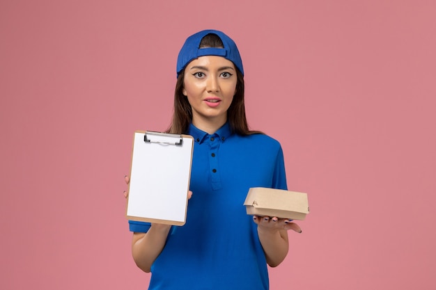 Vue de face femme courrier en cape uniforme bleu tenant peu de colis de livraison avec bloc-notes sur mur rose clair, la livraison des employés de service