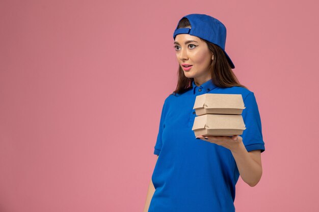 Vue de face femme courrier en cape uniforme bleu tenant peu de colis de livraison sur le mur rose, la prestation de travail de service des employés