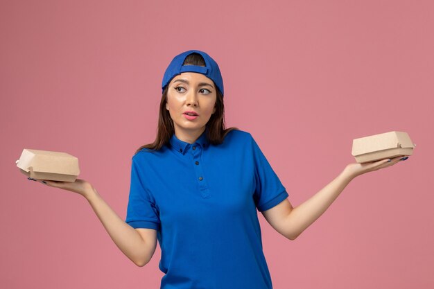 Vue de face femme courrier en cape uniforme bleu tenant peu de colis de livraison sur le mur rose, la prestation de travail de travail des employés