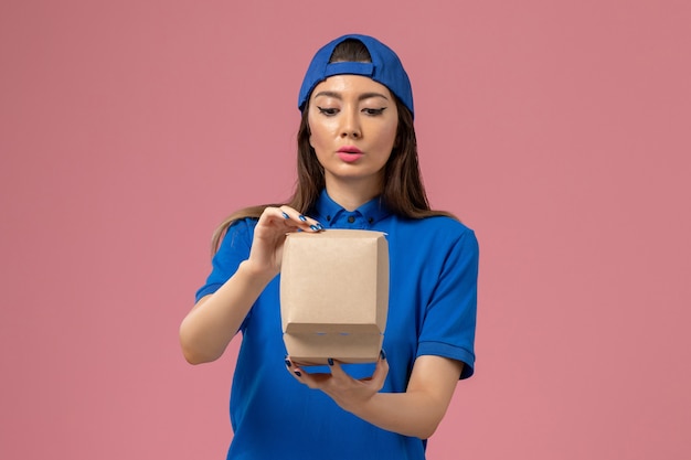 Vue de face femme courrier en cape uniforme bleu tenant peu de colis de livraison et l'ouvrir sur le mur rose, la livraison de l'entreprise de services aux employés